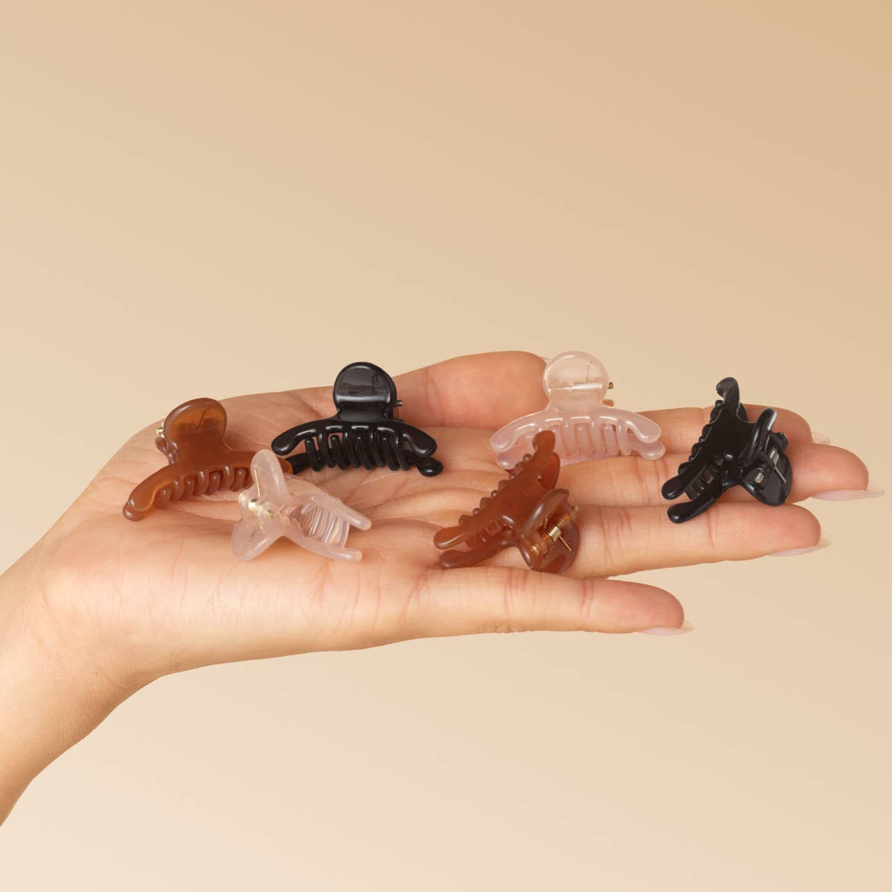 Five small hair claw clips in assorted colors including black, translucent brown, and clear, displayed on an open hand against a solid beige background. Kitsch Arched Clips set of 8 all-day hold hair clips in assorted colors. Pink, brown and black