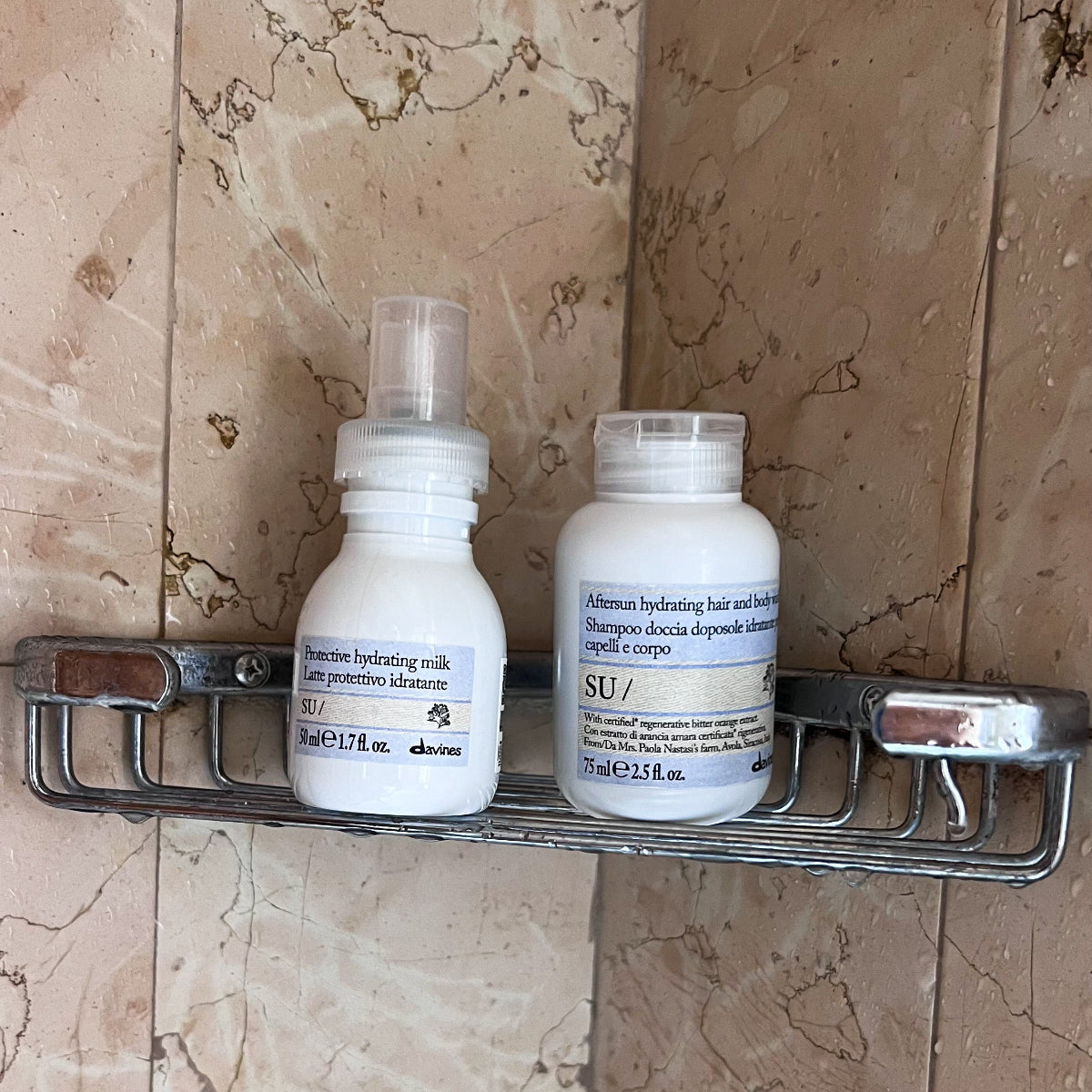 Su Hydrating milk and shampoo and body wash travel bottles from DAVINES SU!, placed on a chrome wall-mounted shelf against beige marble tiles.