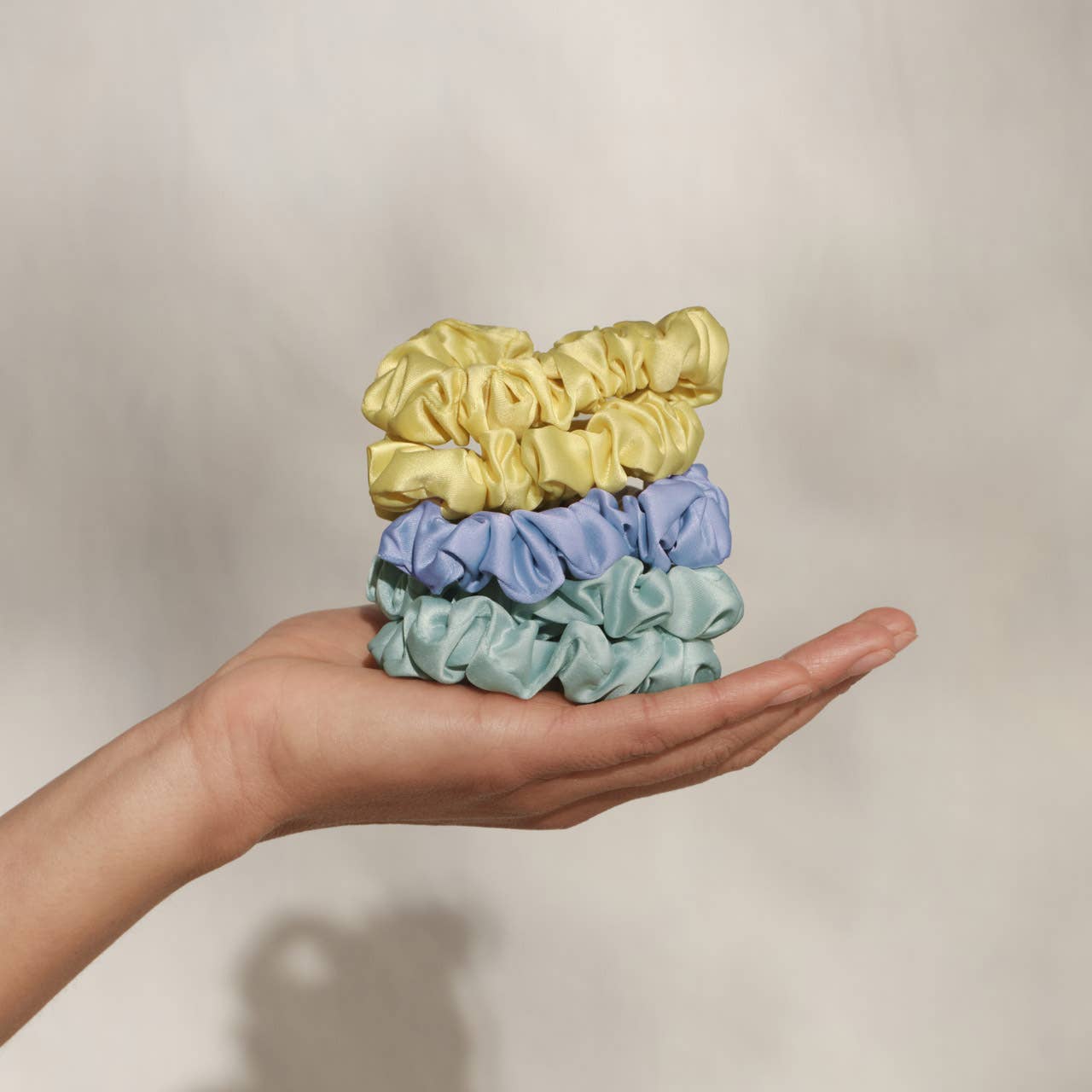 Hand holding three colorful KITSCH scrunchies against a neutral background. Yellow, Blue, Mint Green