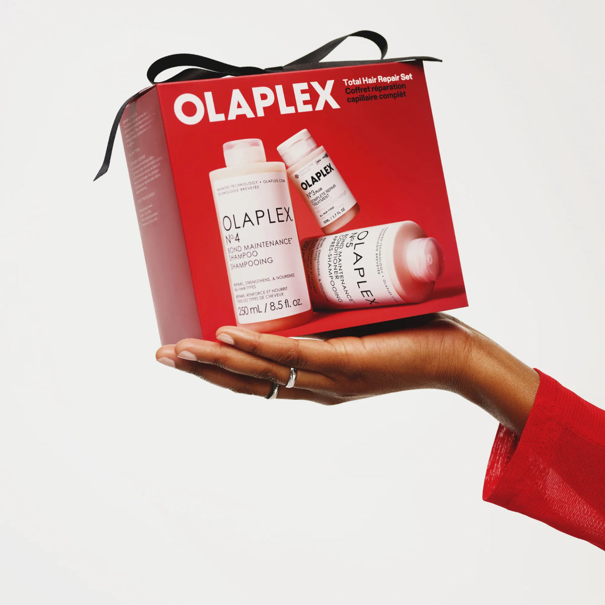A hand holds an Olaplex Total Hair Repair Holiday Set gift box with a black ribbon, featuring the Olaplex No.4, No.3 and No.5