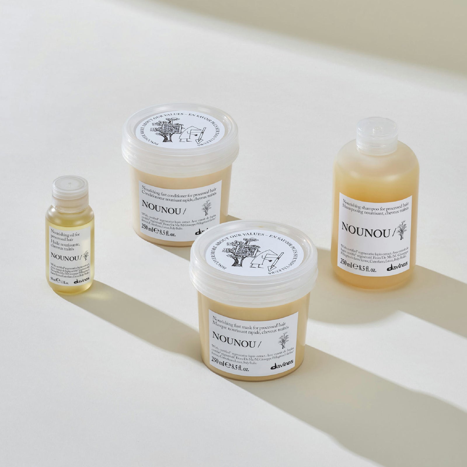 Four NOUNOU skincare products arranged on a clean white surface with soft shadows: a small bottle, a large tub, a medium tub, and a translucent bottle containing pale yellow liquid. The products have white labels with black text and illustrations, and the brand name NOUNOU is clearly visible.