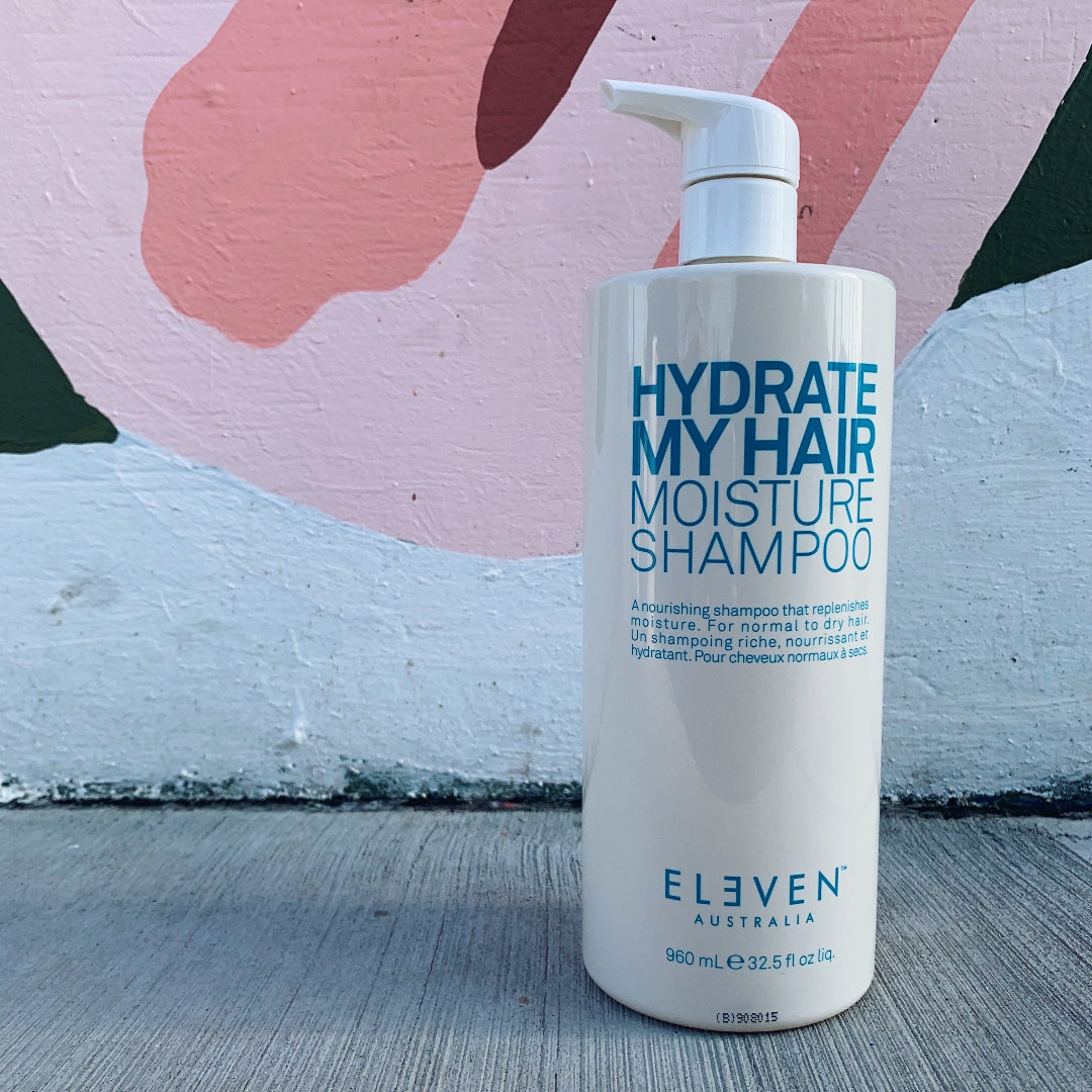 Shampoo bottle labeled 'Hydrate My Hair Moisture Shampoo' by 'Eleven Australia' on a textured surface with abstract art in the background.