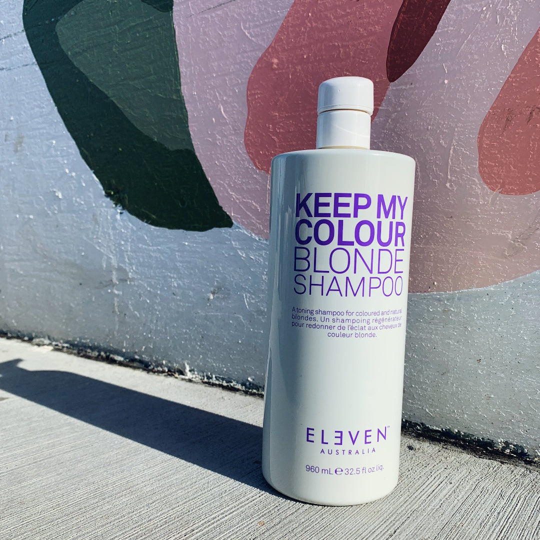 Shampoo bottle labeled 'Keep My Colour Blonde' by Eleven Australia against a graffiti-covered wall.